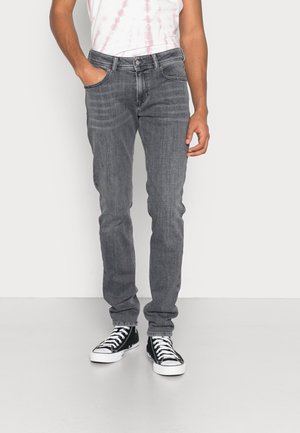 Hombre vistiendo jeans gris ajustados con las manos en los bolsillos y zapatillas negras de caña alta, de pie contra un fondo blanco.