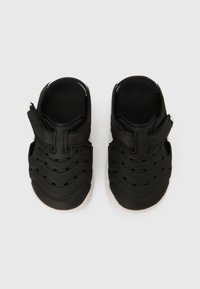Zapatos negros para niños con parte superior de goma que presenta patrones ondulados, lados abiertos y una correa de velcro. La suela blanca contrasta con el material superior.