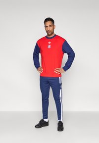 Κόκκινο φούτερ με ναυτικά μανίκια, με κυκλικό λογότυπο και στρογγυλή λαιμόκοψη· συνδυασμένο με ναυτικό παντελόνι με λευκές ρίγες στα πλαϊνά. Μαύρα παπούτσια.