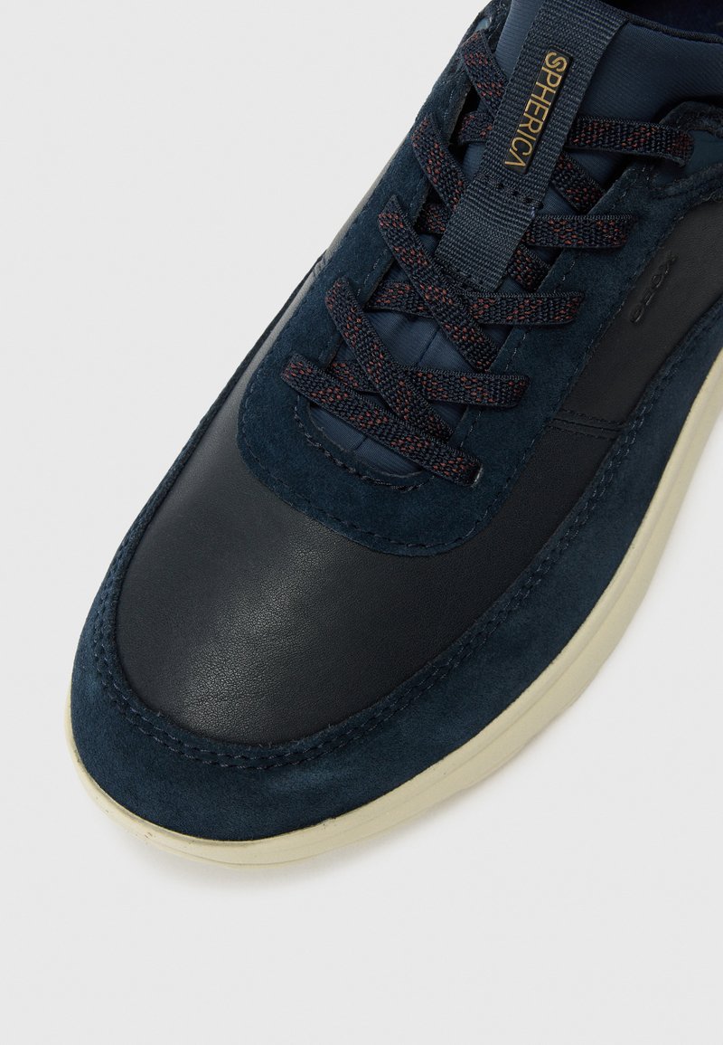 Zapatilla de deporte azul marino que presenta una combinación de cuero liso y suave ante, con cordones texturizados en contraste y una suela de color crema.