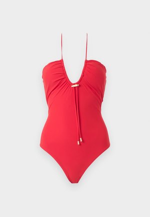 Γυναίκα με βρεγμένα μαλλιά στέκεται ξυπόλυτη, φορώντας ένα κόκκινο ολόσωμο μαγιό με halter-neck, τα χέρια στη μέση, μπροστά σε λευκό φόντο.