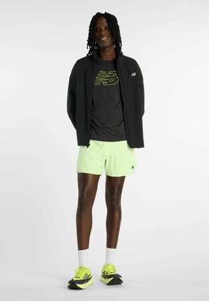 Personne debout souriante, portant une veste noire et un t-shirt avec un logo jaune néon, un short jaune néon, des chaussettes blanches et des baskets assorties.