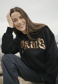 Felpa nera con la scritta "PARIS" in un font a stampa animalier marrone testurizzato, abbinata a jeans grigi scuri. Tessuto liscio, vestibilità rilassata.