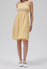 Žluté kostkované šaty s nabíraným živůtkem, tenkými ramínky a rozšířenou sukní. Materiál vypadá lehký a texturovaný, ideální na teplé počasí.