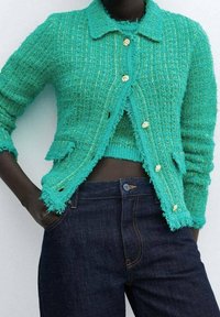 Türkise strukturierte Strickjacke mit Kragen, ausgestattet mit goldfarbenen Knöpfen und Taschen vorne, kombiniert mit dunklen Jeans.