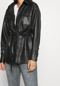 Černá kožená oversize košile-bunda se dvěma kapsami na prsou, manžetami na knoflíky a detailem zavazování v pase, v kombinaci se šedými džínami.