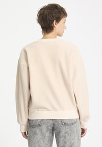 Kremowy polarowy sweatshirt z prążkowanymi ściągaczami i dołem, o luźnym kroju z widocznymi detalami przeszycia. Noszony z jasnoszarymi dżinsami.