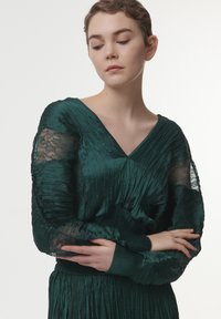 Vestido plisado verde esmeralda con mangas de encaje y escote en V. La tela tiene una textura suave, con detalles fruncidos en la cintura.