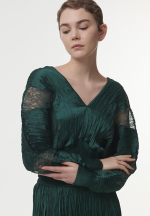 Robe plissée vert émeraude avec des manches en dentelle et un décolleté en V. Le tissu a une texture lisse, avec des détails froncés à la taille.
