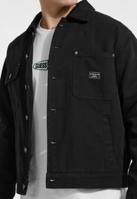 Chaqueta de mezclilla negra con cierres de botón, dos bolsillos frontales y una etiqueta de parche; llevada sobre una camiseta blanca con texto del logo verde.