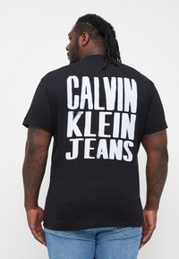 Černé bavlněné tričko s výrazným bílým textem "CALVIN KLEIN JEANS" na zadní straně. Krátké rukávy, běžný střih, měkká textura. Viditelné modré džínové kalhoty.
