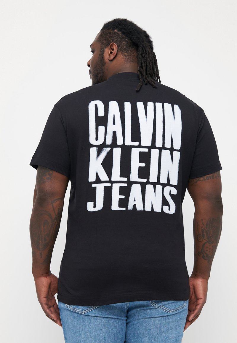 Černé bavlněné tričko s výrazným bílým textem "CALVIN KLEIN JEANS" na zadní straně. Krátké rukávy, běžný střih, měkká textura. Viditelné modré džínové kalhoty.