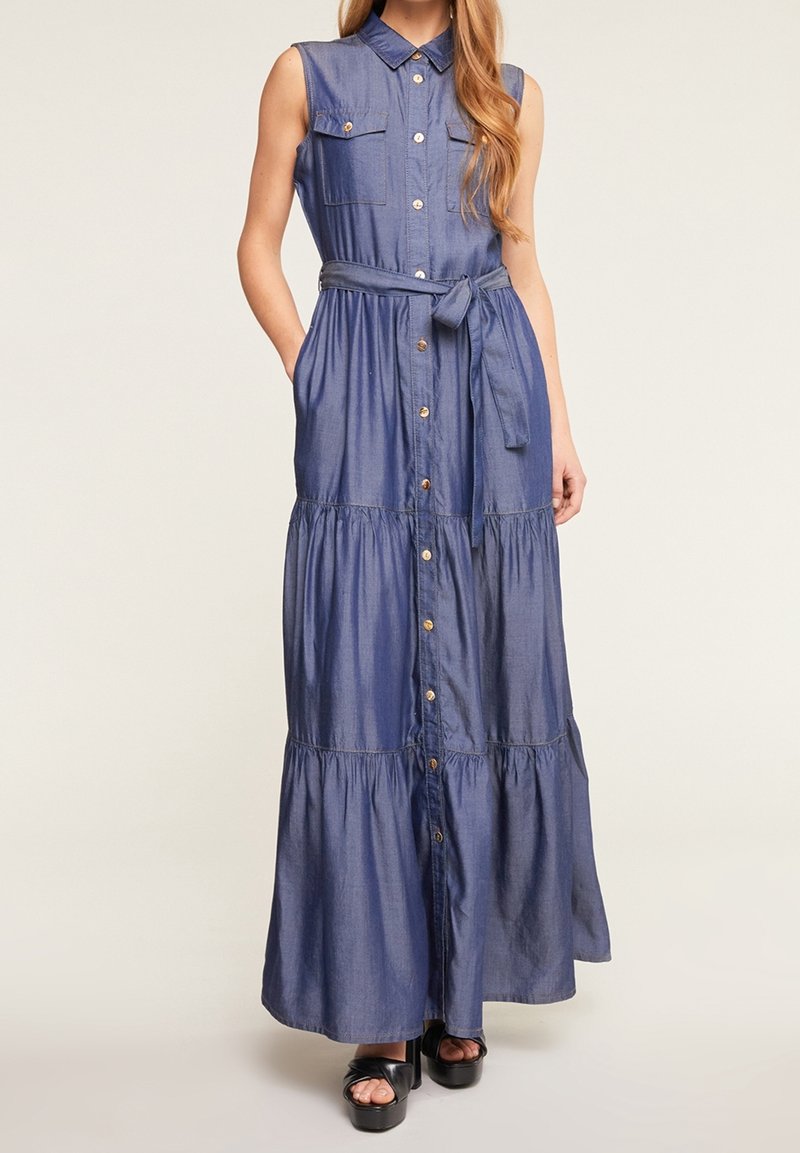 Robe maxi en denim bleu sans manches avec boutons sur le devant, poches poitrine, ceinture à nouer à la taille, portée avec des sandales compensées noires.