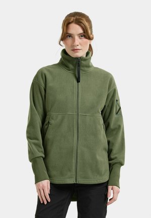 Frau, die eine grüne Fleecejacke mit hohem Kragen, Frontreißverschluss und langen Ärmeln trägt, vor einem neutralen Hintergrund stehend.