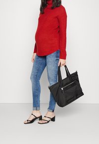 Svart nylon tote-väska med dubbla handtag, frampocket med dragkedja och en slät textur, kombinerad med en röd stickad tröja och blå jeans.