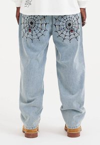 Lichtblauwe spijkerbroek met zwarte spinnenwebborduursels op beide achterzakken en een rode accent, met een relaxte pasvorm en taps toelopende pijpen.