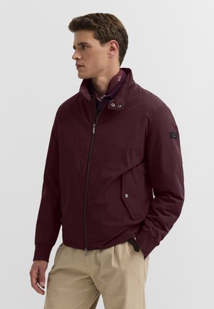 Mann trägt eine weinrote Jacke mit Reißverschluss und geknöpftem Kragen sowie beige Hose, steht mit den Händen in den Taschen und schaut zur Seite.