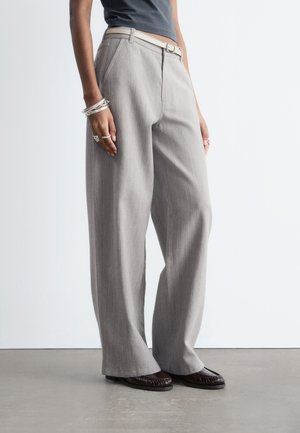 Osoba nosiaca béžové nohavice s vysokým pásom a širokými nohavicami s bielym opaskom, čierny top, strieborné náramky, prstene a tmavohnedé baleríny na sivom podlahe.