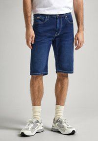 Pantalones cortos de mezclilla azul oscuro con un corte recto, con costuras claras, cinco bolsillos y dobladillo enrollado. Combinados con zapatillas grises y calcetines blancos.