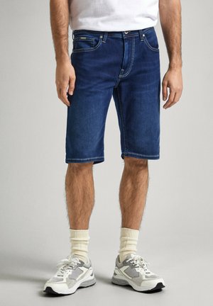 Pantalones cortos de mezclilla azul oscuro con un corte recto, con costuras claras, cinco bolsillos y dobladillo enrollado. Combinados con zapatillas grises y calcetines blancos.