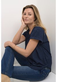 T-shirt à manches courtes bleu marine avec un motif à œillets, associé à un jean bleu taille moyenne. Modèle assis sur un fond blanc.