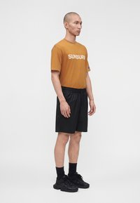 Hombre de pie mirando hacia la derecha, vistiendo una camiseta marrón con la palabra "SUNBURN", pantalones cortos negros, calcetines negros y zapatillas deportivas negras sobre un fondo blanco.