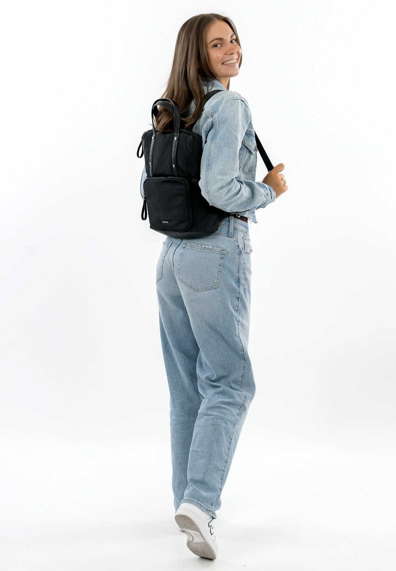 Mochila de tela negra con correas ajustables y asa superior, forma rectangular con un bolsillo frontal con cremallera y detalles en acero inoxidable.