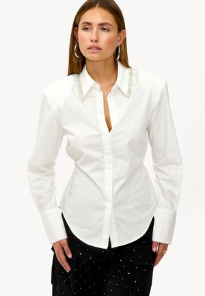 Chemise blanche à boutons, avec un design ajusté, présentant des détails de col embellis et des manches longues. Texture de coton lisse.