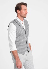 Chaleco a cuadros gris hecho de tela texturizada, de un solo botón con solapas de muesca y botones. Llevado sobre una camisa blanca de cuello.