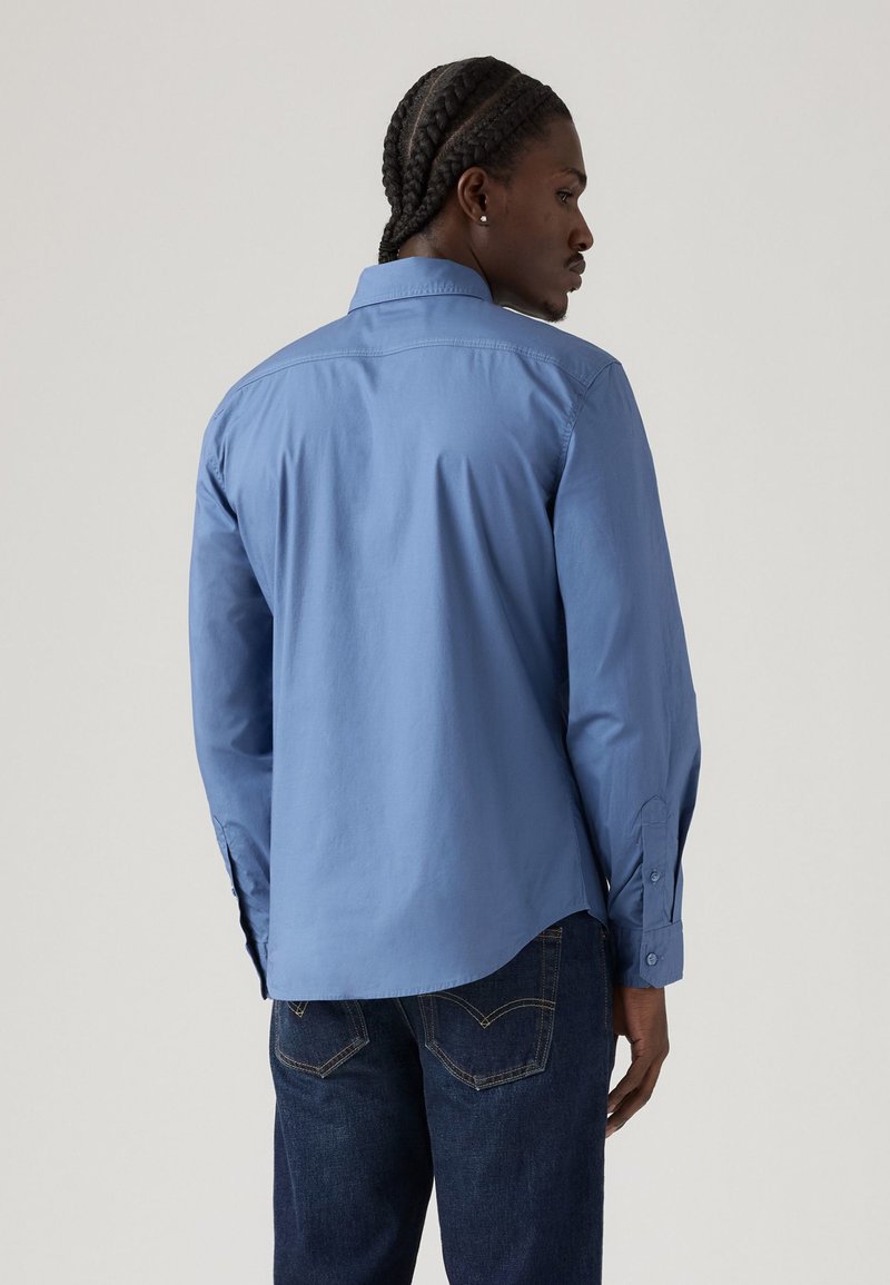 Chemise à manches longues bleu clair avec col pointu, poignets boutonnés et ourlet arrondi. Elle présente une texture lisse semblable au coton et un design minimaliste.