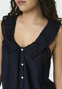Navyblauwe mouwloze top met een gerimpelde, geborduurde kraag, voorzien van knoopsluiting en bloemenpatronen op de stof.