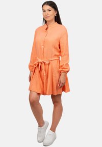 Abito camicia button-up in tessuto arancione testurizzato, con maniche lunghe, una cintura coordinata e una gonna a balze. Indossato con sneakers bianche.