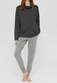 Szary top z kołnierzem w formie cowl i długim rękawem, zestawiony z jasnoszarymi, zwężającymi się spodniami dresowymi, oba wykonane z miękkiego materiału. Na bose stopy, o minimalistycznym designie.