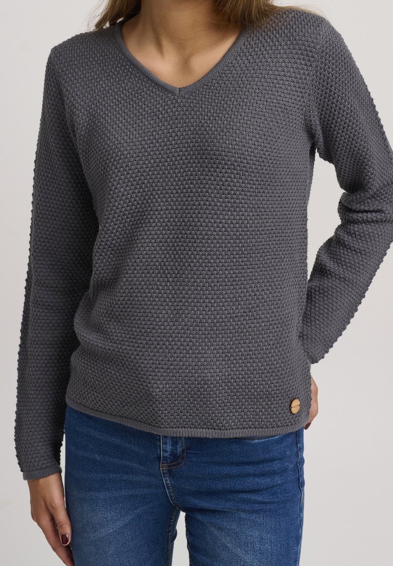 Pull en maille grise avec un col en V, un motif texturé et des manches longues. Présente un petit logo en bois sur l'ourlet. Porté avec un jean en denim.