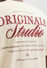 Κρεμ παντελόνι βαμβακερό μπλουζάκι με έντονο μπορντό κείμενο, περιλαμβάνοντας "ORIGINALS" και "Στούντιο", με διακοσμητική γραφή και πρόσθετες φράσεις.