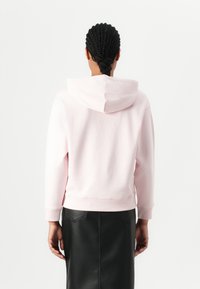 Hoodie rosa chiaro con tasca a marsupio frontale, polsini e orlo a coste. Presenta un cappuccio regolabile con cordino, abbinato a una gonna in pelle nera.