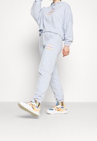 Felpa in cotone azzurro chiaro con un logo grafico, abbinata a pantaloni della tuta coordinati con un dettaglio ricamato, e sneakers bianche con accenti colorati.