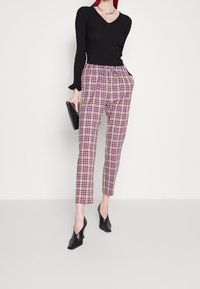 Haut noir à rayures, pantalon à carreaux dans des teintes de violet, rose et beige avec taille élastique, associé à des chaussures à talons noires et une pochette.