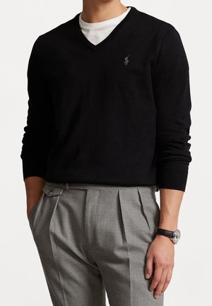 Mand iført sort V-hals sweater over hvid skjorte, grå plisserede bukser og et sølvur med sort læderrem.