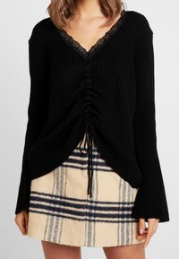 Maglione nero lavorato a maglia con scollo a V e bordo in pizzo, caratterizzato da dettagli plissettati e lacci regolabili, abbinato a una gonna beige con motivo a fantasia.