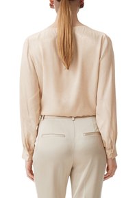 Kvinde med langt, glat hår iført en lys beige satinbluse og matchende skræddersyede bukser, vist fra ryggen mod en hvid baggrund.