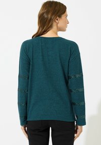 Maglietta a maniche lunghe teal con tessuto morbido, dettagli a strisce riflettenti sulle maniche, scollatura rotonda e un design dal taglio rilassato.