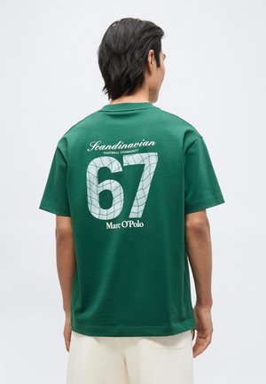 Άνδρας με την πλάτη, φοράει πράσινη μπλούζα με το κείμενο «Scandinavian Football Community 67 Marc O’Polo» στην πλάτη, συνδυασμένη με ανοιχτόχρωμα σορτς.