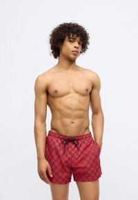 Red swim shorts featuring a repeating pattern in black and pink, with an elastic waistband and a drawstring detail.