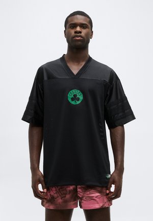 Férfi fekete Celtics mezben és piros mintás rövidnadrágban, fehér, egyszínű háttér előtt, előre nézve áll.