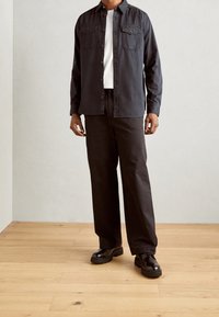 Dunkles Hemd mit Knöpfen und zwei Brusttaschen, über einem weißen T-Shirt getragen. Schwarze Stoffhose und robuste schwarze Schuhe, auf einem Holzboden. 