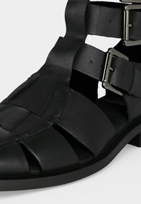 Μαύρο δερμάτινο σανδάλι με δύο ασημένια λουριά με πόρπες και φαρδιές ανοικτές περιοχές στα πλάγια και μπροστά για αερισμό.