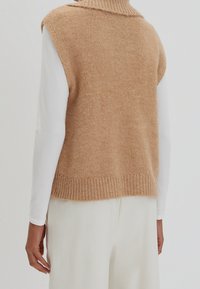 Vue de dos d'une personne portant un gilet sans manches en tricot beige sur un haut blanc à manches longues avec un pantalon blanc ample.