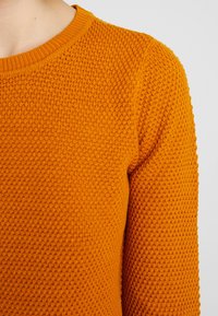 Närbild av en person som bär en texturerad, orange stickad tröja med rund halsringning mot en enfärgad ljus bakgrund.