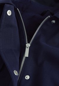 Silberner Metallreißverschluss und Druckknöpfe an teilweise geöffnetem dunkelblauem Stoff, zeigen Details des Verschlussmechanismus der Jacke.